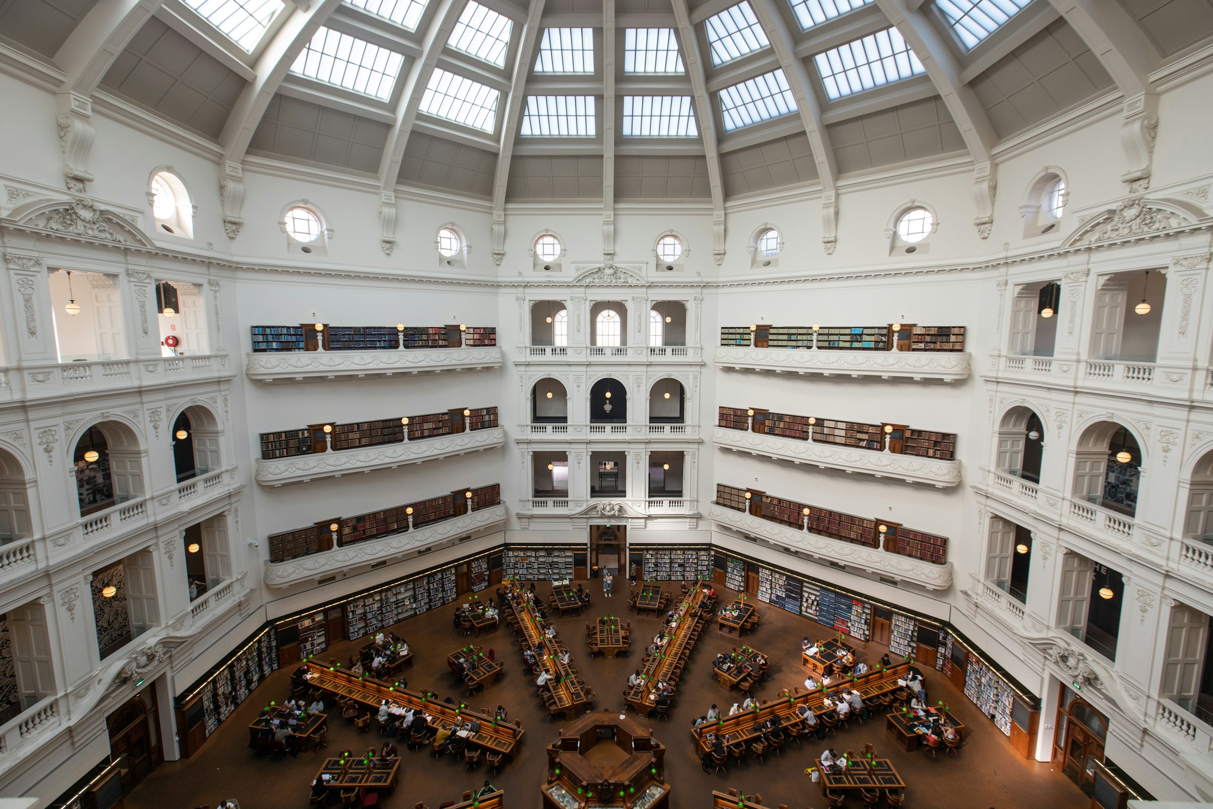 State library of Victoria A photo of a hexagonal building with a glass arched ceiling. It's a library with wooden desks in a start shape. It looks peaceful and beautiful.